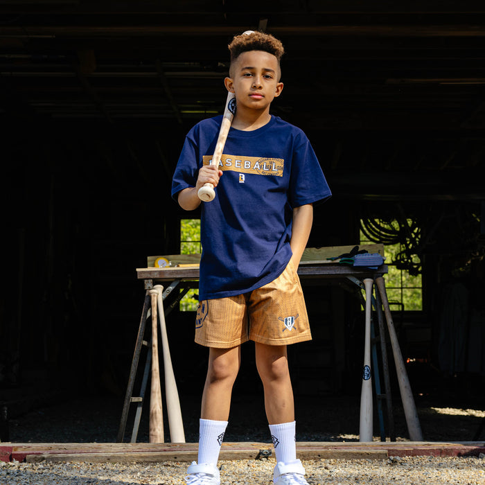Young boy standing confidently with a baseball bat resting on his shoulder wearing the navy youth Radar Tee paired with tan Father & Son Bat Company shorts and white BL101 crew socks