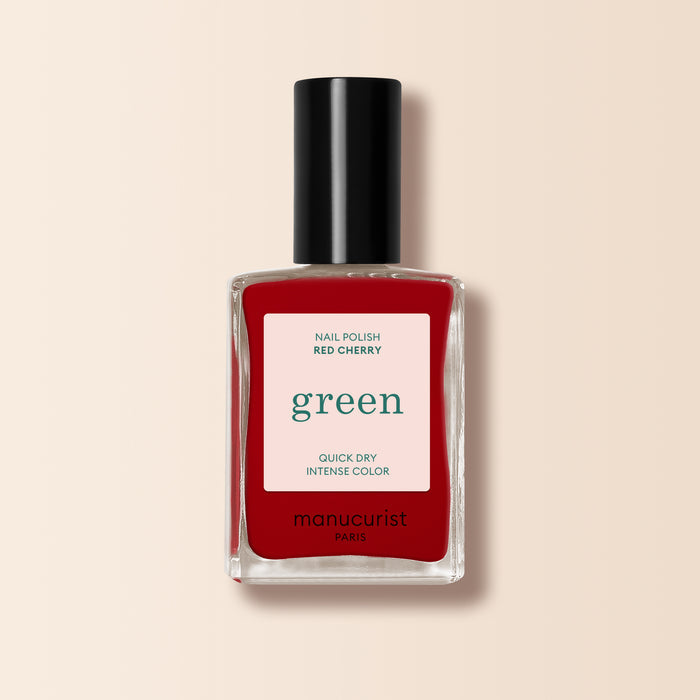 A bottle of Manucurist Red Cherry nail polish with a black cap and "green, quick dry intense color" label is set against a beige background.