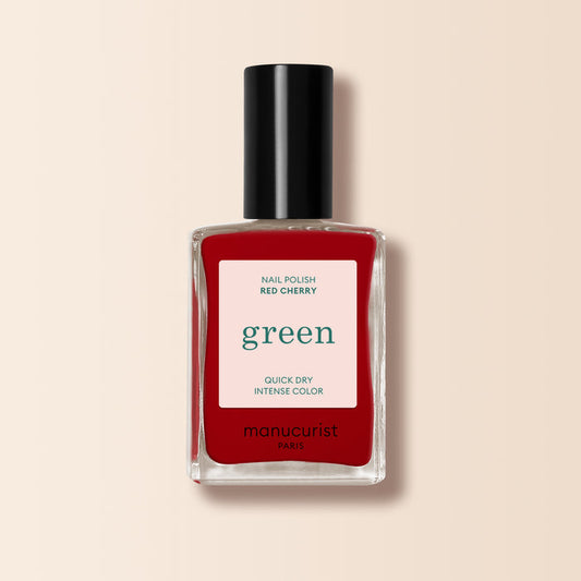 A bottle of Manucurist Red Cherry nail polish with a black cap and "green, quick dry intense color" label is set against a beige background.