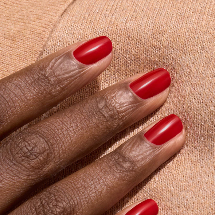 A close-up of a hand with dark skin and neat nails painted in Manucurist's Red Cherry, resting on soft, light beige knit fabric.
