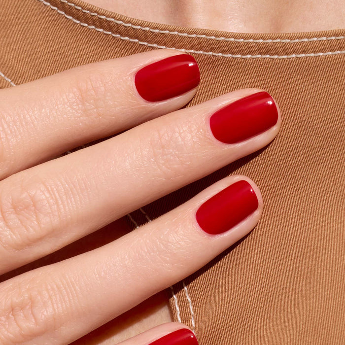 A close-up of a hand with neatly manicured nails painted in Manucurist’s Red Cherry polish, held against tan fabric with white stitching.