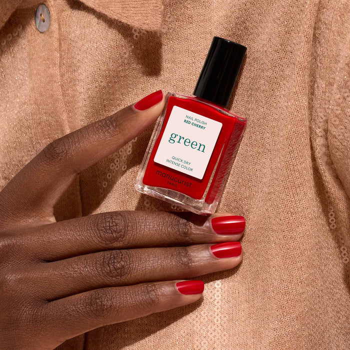 A hand with neatly manicured red nails holds a bottle of Manucurist Red Cherry nail polish against a beige, textured fabric background.