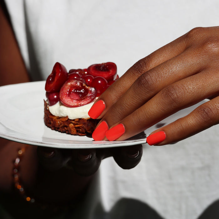 A hand with Manucurist's Red Coral nail polish holds a white plate with a small dessert topped with whipped cream and halved cherries. The person wears a white top and a gold bracelet.