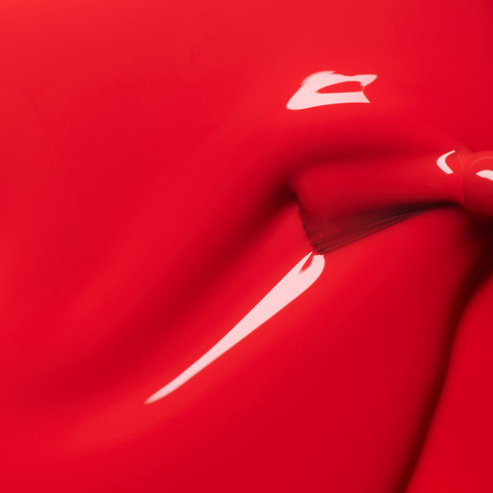 A close-up of glossy Red Coral nail polish from Manucurist being applied with a brush, revealing its smooth texture, shiny surface, and striking light reflections.