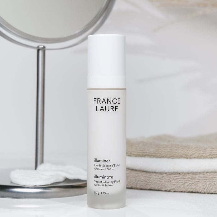 France Laure Illuminate Secret Glowing Fluid with Orchid & Saffron, 50g bottle on vanity beside mirror and towels