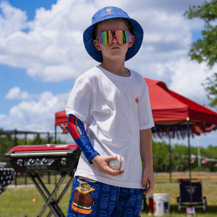 Child wearing a blue bucket hat and colorful sunglasses, holding a baseball, with a red tent and green trees in the background.