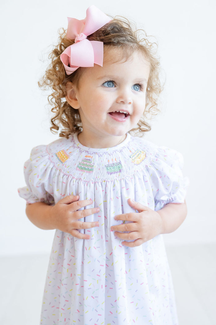 The dress is hanging up on a hanger, showcasing its colorful sprinkle pattern and heirloom design, perfect for babies, toddlers, and young girls.