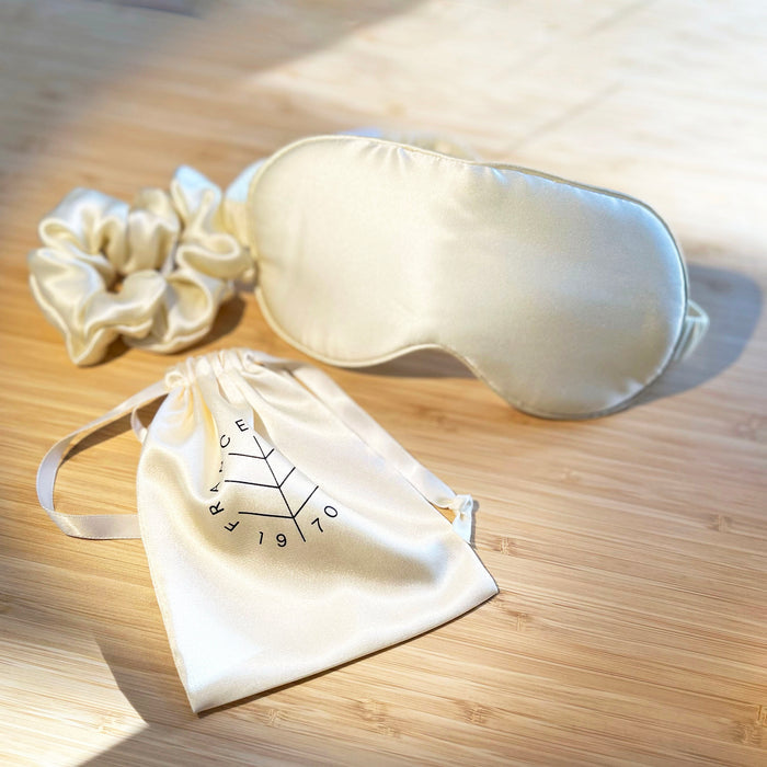 Cream silk sleep mask with matching scrunchie and drawstring pouch, featuring 'FRANCE 1970' logo, displayed on wood surface.