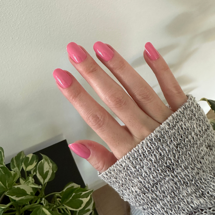 Taffy | Short | Round - Tab Press-On showing glossy bright pink press-on nails on hand with cozy sweater sleeve and plants background