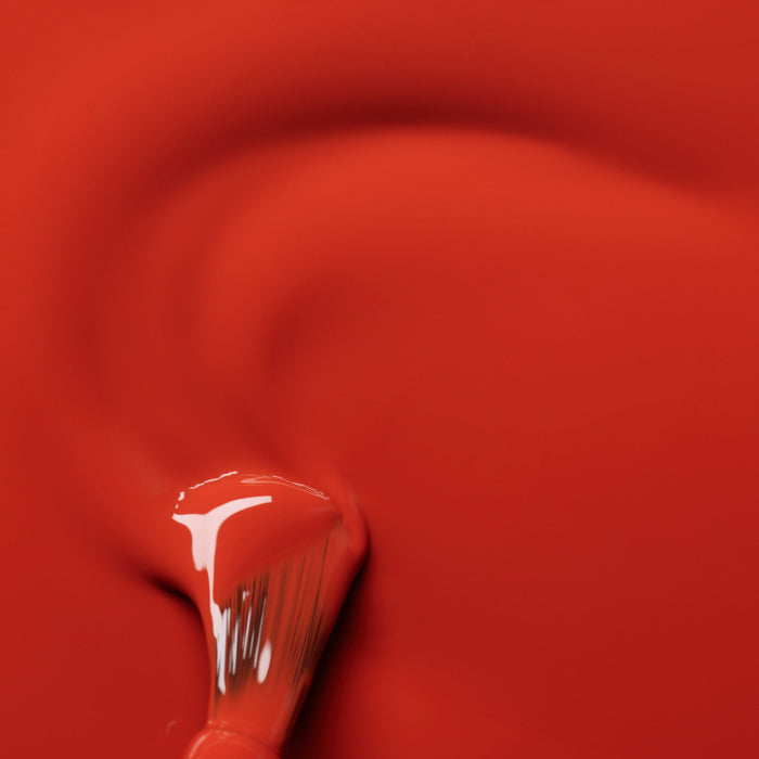 A close-up of a paintbrush sweeping through glossy, wet Manucurist Terracotta nail polish in scorched red earth, creating smooth curved lines and textures.