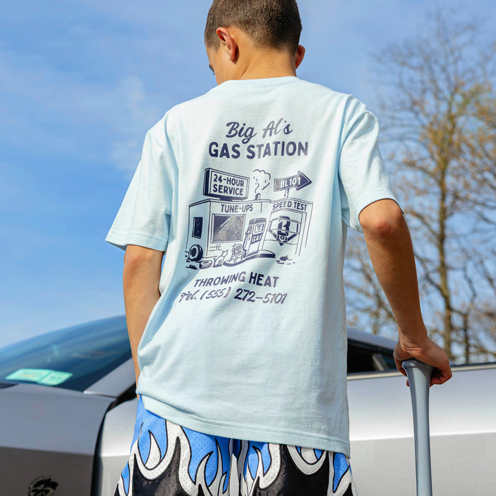 Back of light blue youth tee with navy vintage gas station illustration and "Throwing Heat" text, outdoor setting.