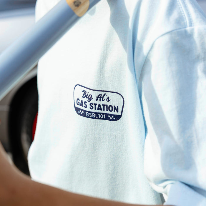 Close-up of navy Big Al's Gas Station BSBL 101 badge graphic on light blue youth tee, bat handle visible at top.