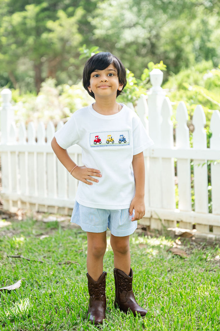 Boy wearing Smocked Tractor T-Shirt and Short Set by Ruth and Ralph, standing outdoors in front of a white picket fence