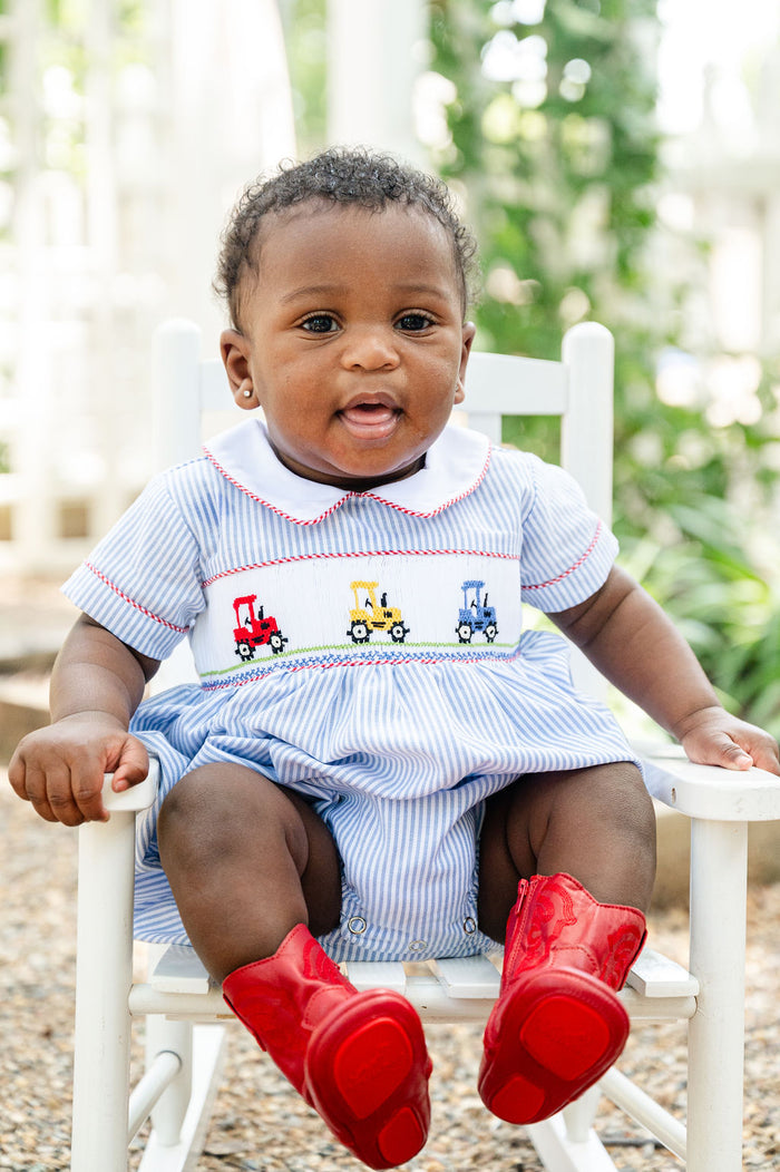 Baby boy wearing Smocked Tractor Bubble by Ruth and Ralph, sitting in white rocking chair outdoors with red boots