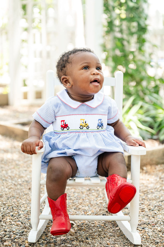Baby boy standing in white chair wearing Smocked Tractor Bubble by Ruth and Ralph with red boots