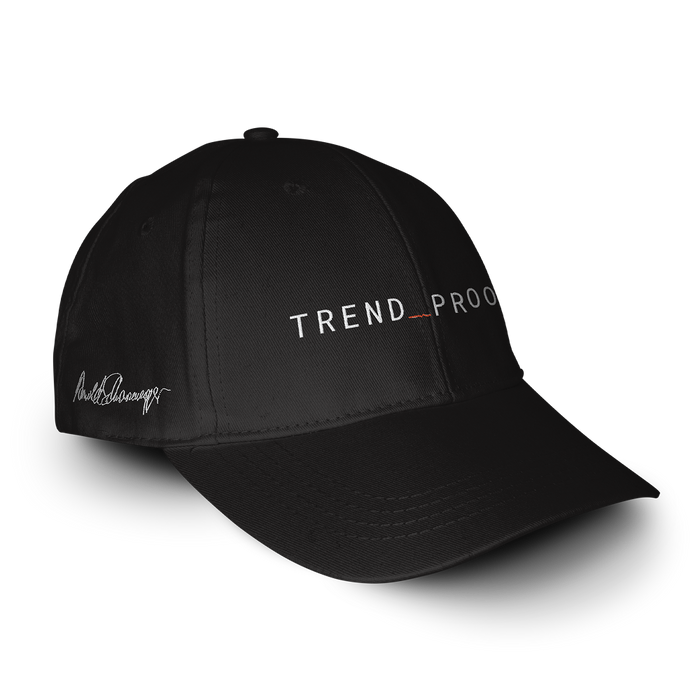 Black “TREND_PROOF” baseball cap shown at an angle, with white embroidered logo on the front and Arnold Schwarzenegger’s signature stitched on the side panel.