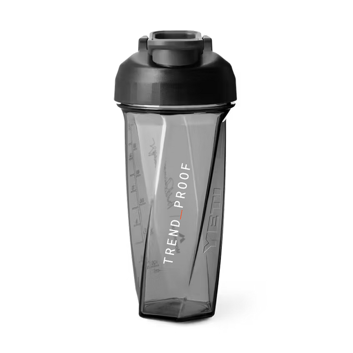 Front view of a transparent gray Helimix shaker bottle with a black lid and flip cap. The bottle has an angular, faceted design with measurement markings along the side and the word “HELIMIX” embossed vertically on the right side panel. Printed vertically on the front in white text is “TREND_PROOF” with an orange underscore between the words. The shaker is designed for clean mixing and everyday training use.