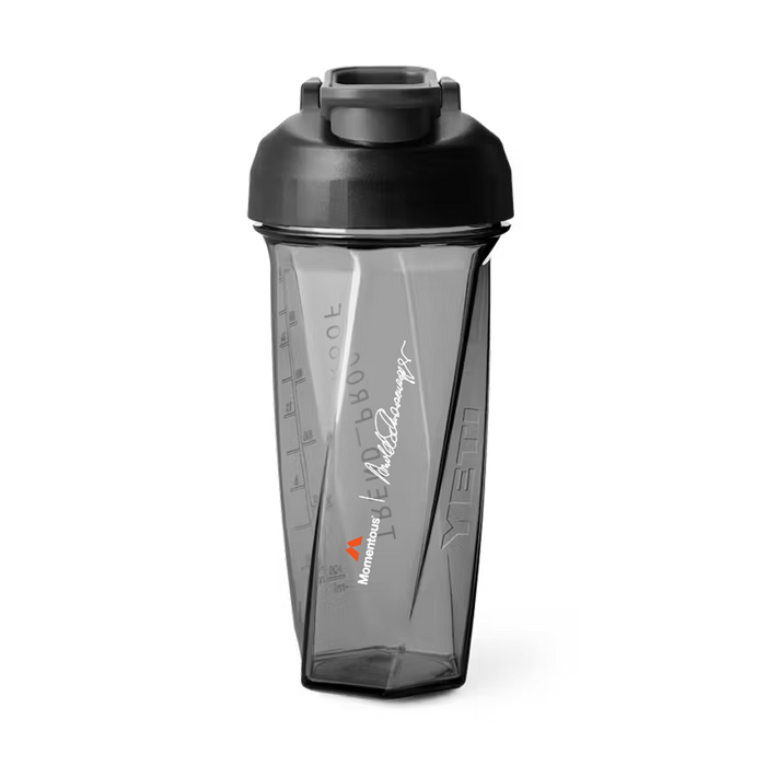 Front view of a transparent gray Helimix shaker bottle with a black screw-top lid and flip cap. The bottle has a faceted, geometric shape with embossed measurement markings on the side. Printed vertically on the front is a white Arnold Schwarzenegger signature above a small orange Momentous logo and white “Momentous” wordmark. The bottle appears designed for mixing supplements or protein shakes.