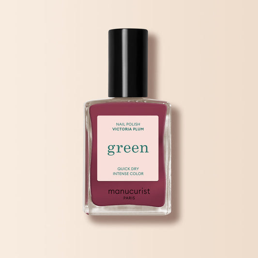A bottle of Manucurist Victoria Plum nail polish, featuring a deep plum shade, black cap, and white label with green and pink text, is displayed on a light beige background.