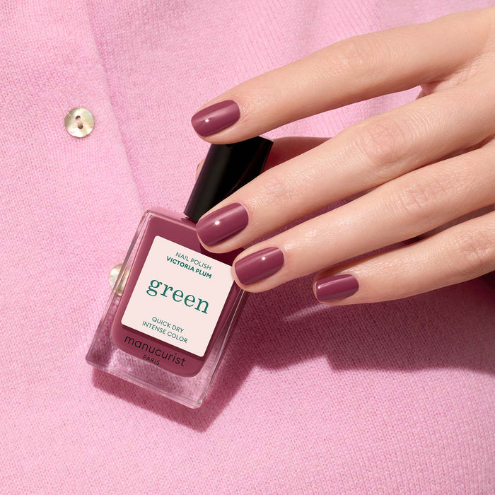 A hand with mauve nails holds a bottle of Manucurist's Victoria Plum nail polish against a light pink fabric background.